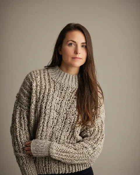 A woman in a chunky beige knit sweater stands against a gray background.