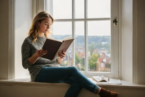 Young woman reading a book on a windowsill with city view